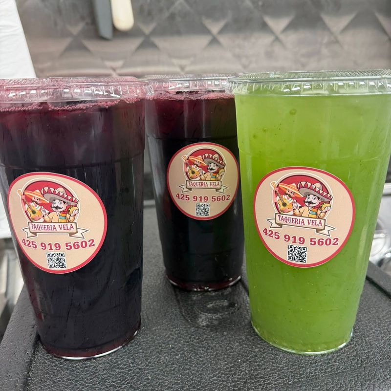 Three cups of aguas frescas with Taquería Vela stickers, including jamaica and pepino con limón